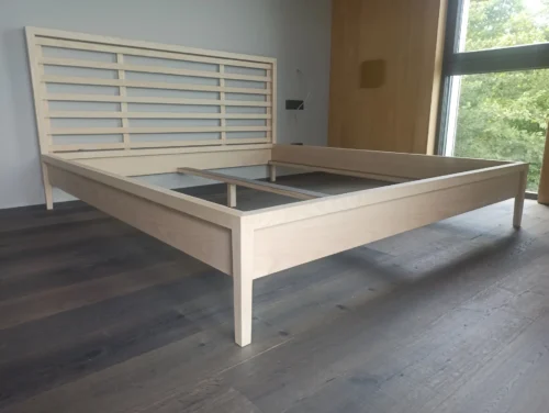 Freistehendes Bettgestell aus hellem Eschenholz. Das Kopfteil besteht aus einer Rahmenkonstruktion mit horizontal verlaufenden Holzleisten. Die Konstruktion zeigt schlanke, konisch zulaufende Bettpfosten und eine mittige Stützleiste für den Lattenrost.