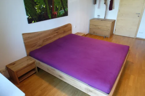 Massivholz-Doppelbett aus Eiche mit einem organisch geschwungenen Kopfteil. Das Set umfasst zwei Nachtkästchen mit jeweils einer Schublade. Im Hintergrund ist eine passende Kommode sichtbar.