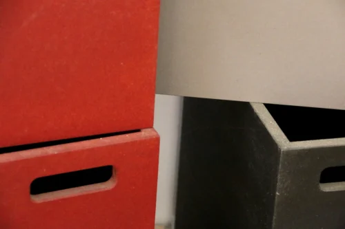 Detailaufnahme von ineinandergreifenden, maßgefertigten Korpus-Elementen aus durchgefärbtem Holzwerkstoff in den Farben Rot, Anthrazit und Grau. Sichtbar sind präzise gefräste Griffmulden und exakte Kantenführungen.