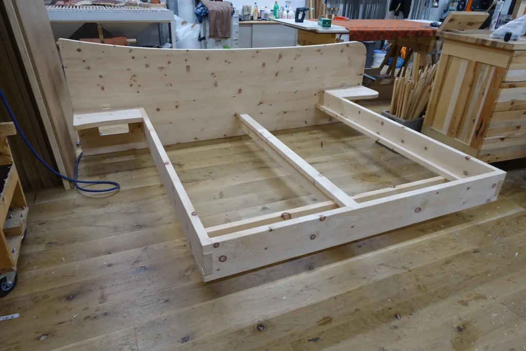 Großformatiges Familienbett aus massivem, astreichem Zirbenholz im Rohzustand in der Werkstatt. Das Bett verfügt über ein hohes, geschwungenes Kopfteil mit zwei integrierten, schwebenden Nachtablagen. Die stabile Rahmenkonstruktion zeigt eine mittige Längstraverse und zwei Querverstrebungen zur Aufnahme der Lattenroste.