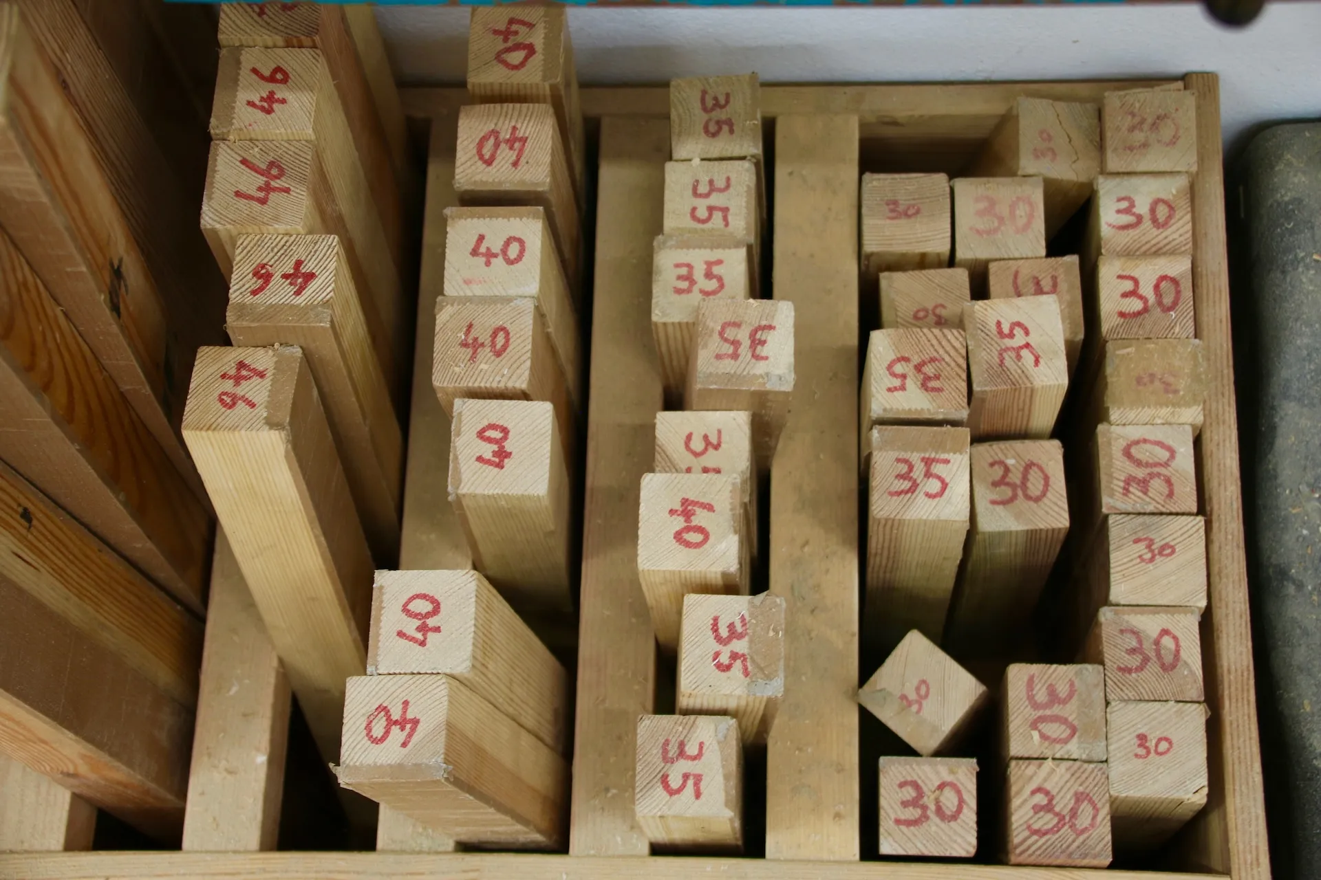 26 Werkstatt und Handwerk 14 Vogelperspektive auf ein hölzernes Sortierregal in der Werkstatt, in dem zahlreiche quadratische Massivholz-Leisten stehend gelagert werden. Die Stirnflächen der Hölzer sind mit roten Zahlen (z.B. 30, 35, 40, 46) markiert, die die jeweilige Länge oder Stärke kennzeichnen.