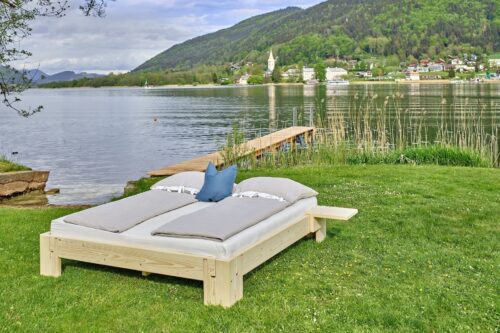 Metallfreies Massivholzbett steht auf einer Wiese direkt am Ossiacher See. Im Hintergrund am anderen Ufer das Stift Ossiach