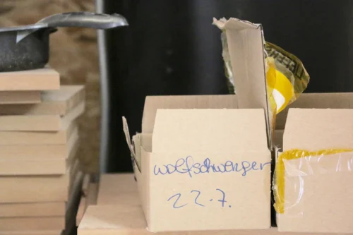 Nahaufnahme eines geöffneten Versandkartons in der Werkstatt, der handschriftlich mit dem Namen 'Wolfschwenger' und einem Datum markiert ist.