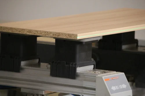 Detailaufnahme einer CNC-Bearbeitung: Eine helle Möbelplatte, auf einer Seite bekantet, ist auf schwarzen Vakuumsaugern fixiert. Ein grüner Positionierlaser projiziert eine exakte Führungslinie auf die Kante des Materials.