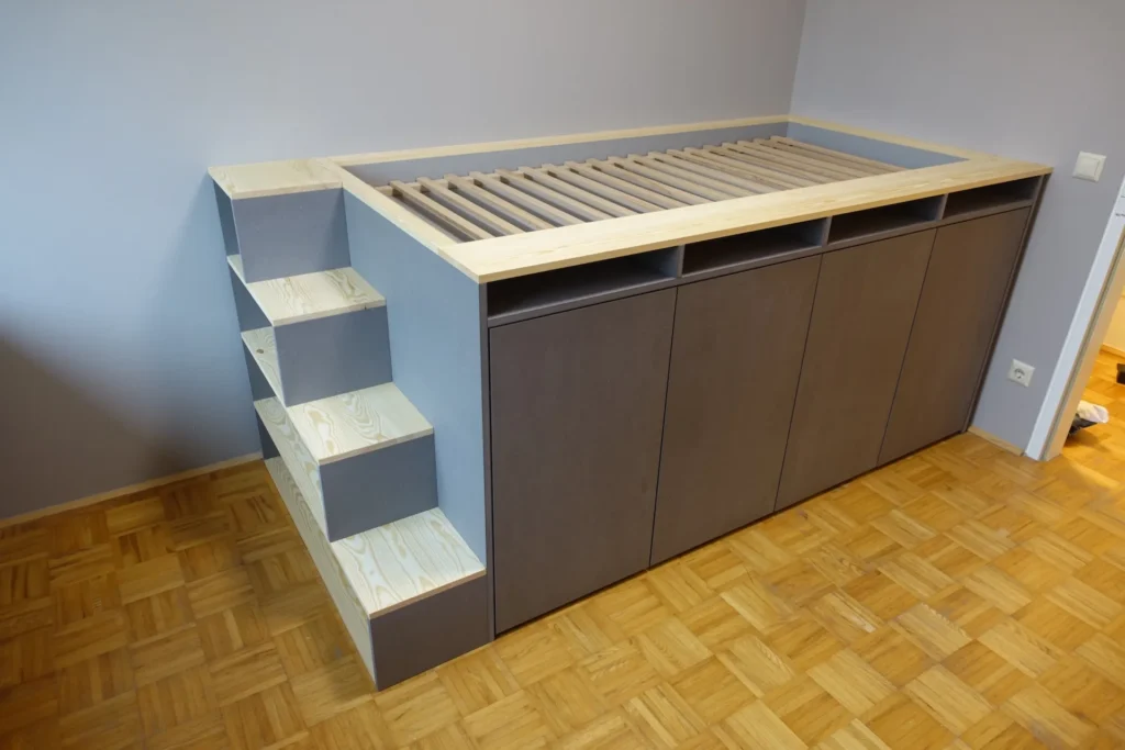 Erhöhtes Podestbett mit integriertem Stauraumsystem. Der Korpus ist in mattem Grau gehalten, während die Trittstufen der seitlichen Treppe und die Bettumrandung aus hellem Massivholz mit ausgeprägter Maserung gefertigt sind.