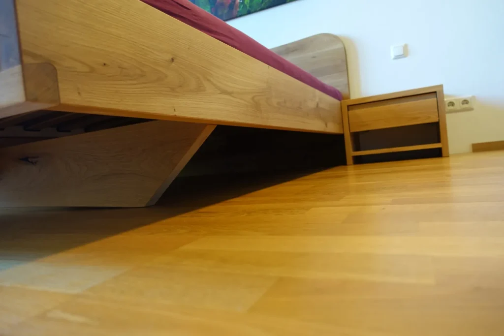 Detailaufnahme eines Refluxbettes aus massivem Eichenholz von der Bodenperspektive aus. Sichtbar ist die keilförmige Unterkonstruktion (Kufen), die den gesamten Bettrahmen in eine therapeutische Schräglage versetzt. Das Bett verfügt über ein passendes Nachtkästchen und steht auf einem hellen Parkettboden; die Liegefläche ist mit einem roten Spannlaken bezogen.