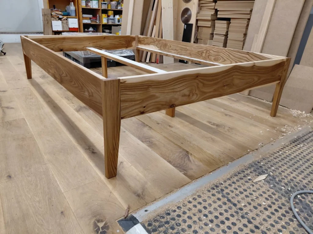 Detailaufnahme eines freistehenden Doppelbettrahmens aus massivem Rüsterholz (Ulme) in der Werkstatt. Die Konstruktion folgt einer Modulbauweise mit metallfreien Steckverbindungen an den Eckpfosten. Die markante, lebhafte Maserung des Ulmenholzes zieht sich über die breiten Zargen.