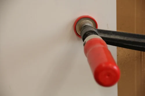 Detailaufnahme einer schwarzen Schraubzwinge mit rotem Griff, die eine weiße, beschichtete Möbelplatte fixiert. Der rote Druckteller der Zwinge setzt präzise auf der hellen Oberfläche an. Das Bild zeigt einen Ausschnitt aus der Montage eines modernen Möbelteils.