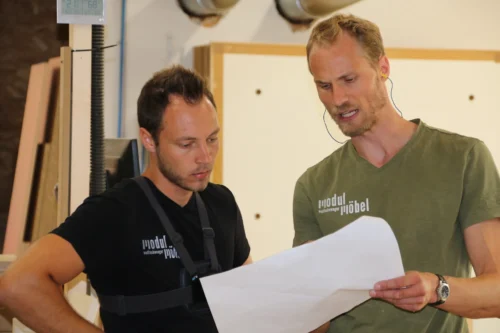 Unsere Mitarbeiter bei der gemeinsamen Projektbeschsprechung. Beide tragen Firmen-T-Shirts. Sie halten einen großen Plan in den Händen und diskutieren konzentriert über die Details.