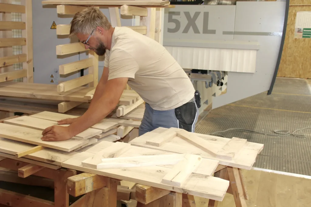 David bei der manuellen Qualitätskontrolle und Sortierung von Massivholz-Elementen auf einem Holzbock in der Werkstatt. Im Hintergrund ist unsere modernes 5-Achs-CNC-Bearbeitungszentrum (Modell 5XL) sowie ein hölzernes Lagerregal für Werkstücke sichtbar.