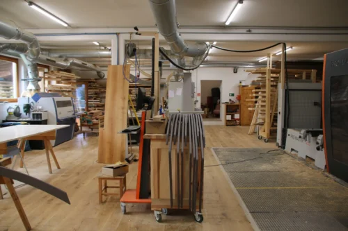 Weitwinkelaufnahme unserer hellen Werkstatt mit Massivholzboden. Im Zentrum stehen auf einem Rollwagen mehrere gebogene, anthrazitfarbene Formteile. Rechts befindet sich ein modernes CNC-Bearbeitungszentrum (HOLZ-HER PRO-MASTER), links eine Hobelmaschine (Weinig Unimat). Über den Maschinen verläuft ein verzweigtes System aus metallischen Absaugrohren.