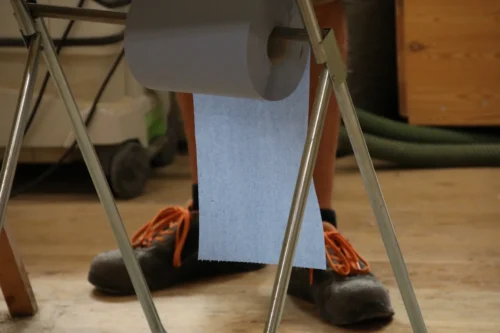 Detailaufnahme einer blauen Putztuchrolle aus Papier auf einem Metallständer in der Werkstatt. Im unscharfen Hintergrund sind die Beine eines Handwerkers in Arbeitsschuhen mit auffälligen orangefarbenen Schnürsenkeln sowie ein mobiler Werkstatt-Absauger erkennbar.
